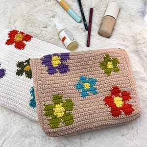 May include: Two crochet cosmetic bags. One is white with red, green, and blue flower designs. The other is light pink with purple, green, blue, and red flower designs. Makeup items are scattered around the bags.