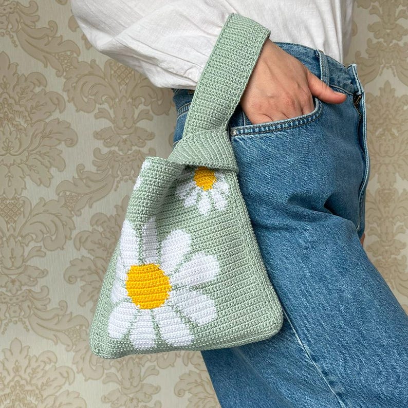 May include: A light green crocheted bag with a white daisy pattern. The bag has a knotted handle and is made with a soft, textured yarn.