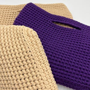 May include: Close-up of three handmade crochet bags. One bag is a deep purple color, featuring a handle opening. Two bags are a light beige color. The bags are made with a thick yarn, showcasing a textured stitch pattern.