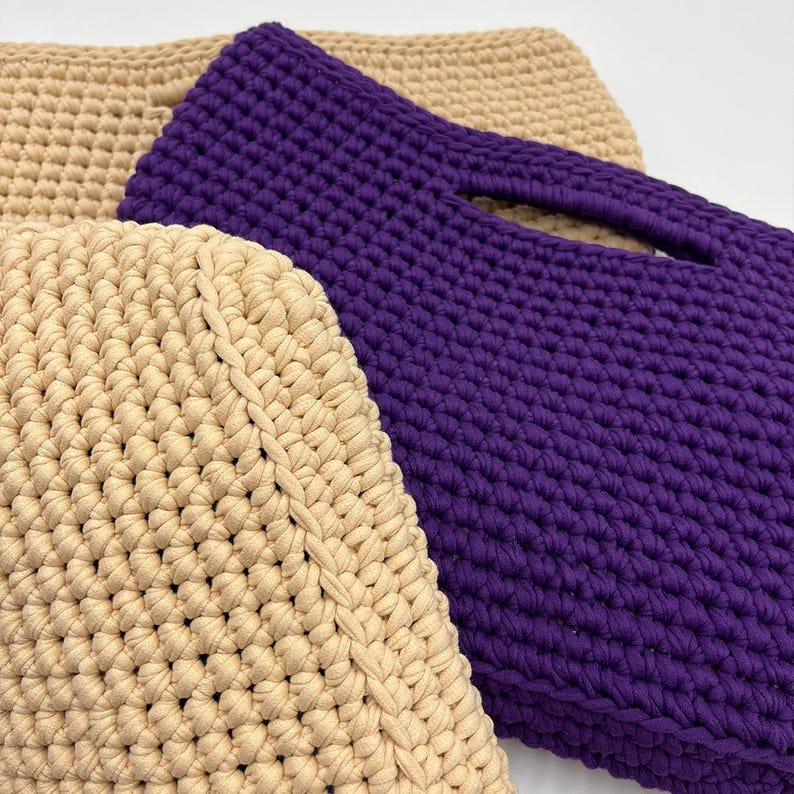 May include: Close-up of three handmade crochet bags. One bag is a deep purple color, featuring a handle opening. Two bags are a light beige color. The bags are made with a thick yarn, showcasing a textured stitch pattern.