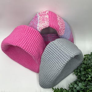 May include: Three crocheted hats in shades of pink and gray. One hat is solid bright pink, another is gray, and the third is striped with pink, white, and lavender. The hats are displayed on a white surface.