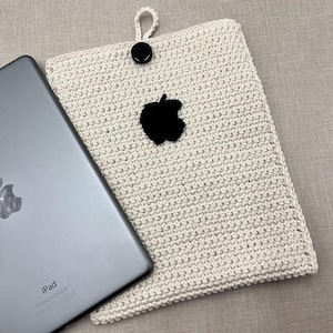 May include: A white crocheted iPad case with a black apple logo. The case is made with a textured stitch and has a simple design.