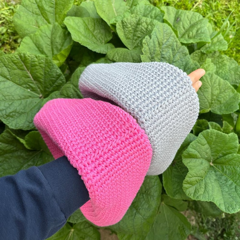 May include: A pair of handmade crochet hats in bright pink and light gray. The hats are made of a thick yarn and have a ribbed texture. The hats are shown against a background of large green leaves.