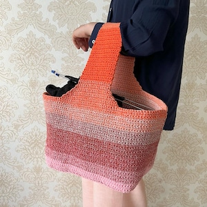 May include: A crocheted tote bag in a gradient of colors from pink to orange. The bag has a rectangular shape with a wide handle. The bag appears to be holding knitting needles and other craft supplies. The bag is held by a person.