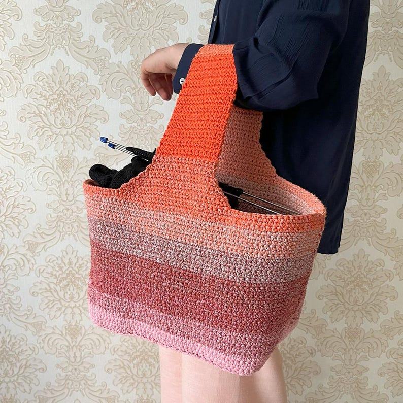 May include: A crocheted tote bag in a gradient of colors from pink to orange. The bag has a rectangular shape with a wide handle. The bag appears to be holding knitting needles and other craft supplies. The bag is held by a person.