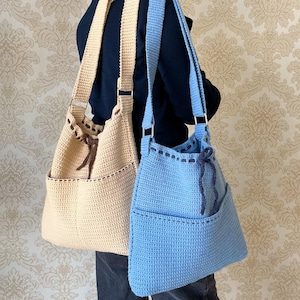 Puede incluir: Dos bolsos de mano de crochet, uno beige y otro azul claro, con asas marrones y cierre de cordón marrón. Los bolsos tienen un bolsillo en la parte delantera.