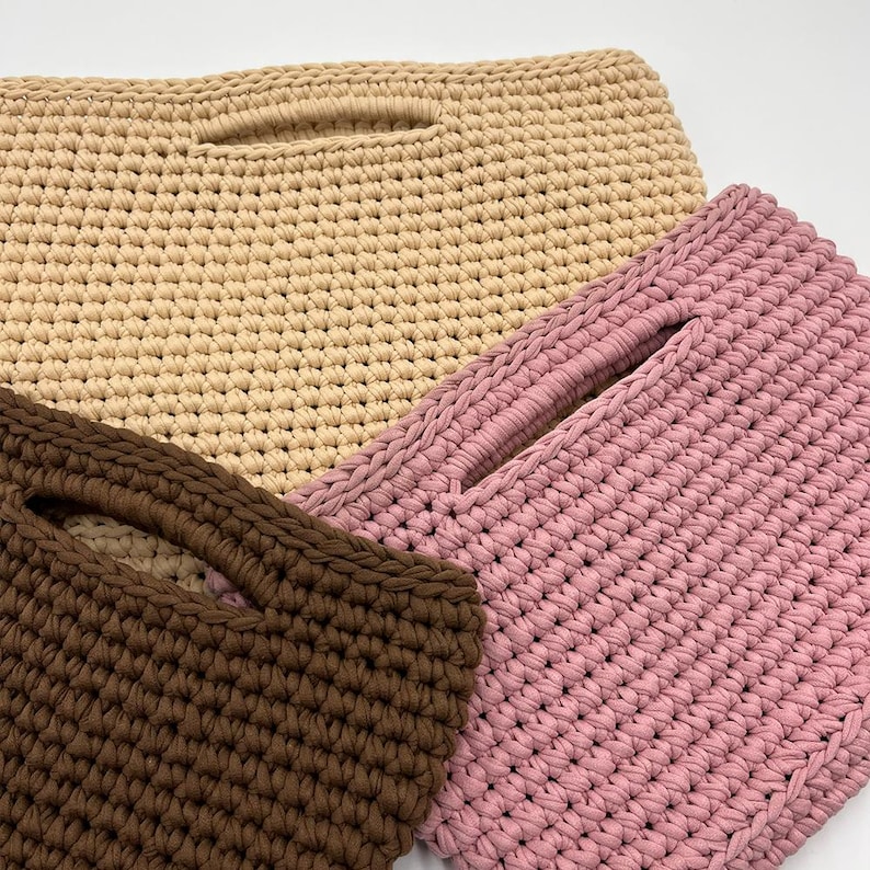 May include: Three handmade crochet bags in varying colors: beige, pink, and brown. Each bag features a rectangular shape with a handle opening. The bags are made with a thick, textured yarn, showcasing the detailed crochet work.