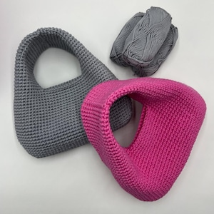 May include: Two handmade crochet bags, one gray and one hot pink, are displayed on a white surface. A ball of gray yarn sits next to the bags. The bags have a rounded shape with a handle.