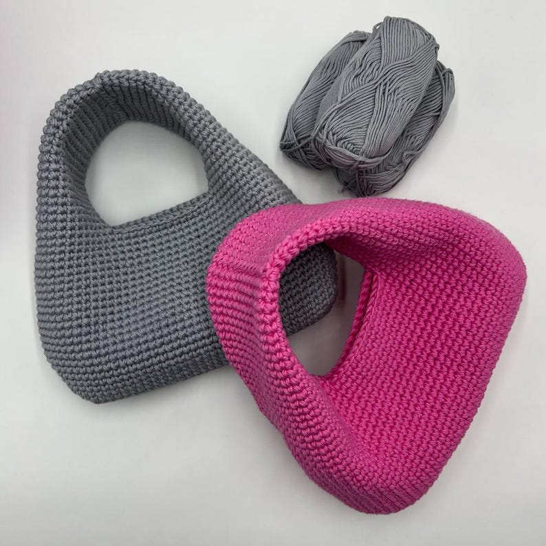 May include: Two handmade crochet bags, one gray and one hot pink, are displayed on a white surface. A ball of gray yarn sits next to the bags. The bags have a rounded shape with a handle.