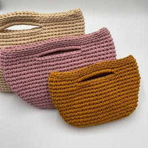 May include: Three handmade crochet bags in beige, pink, and mustard yellow. Each bag features a rectangular shape with a handle opening. The bags are made of thick yarn, showcasing a textured, woven appearance. The bags are displayed on a white surface.