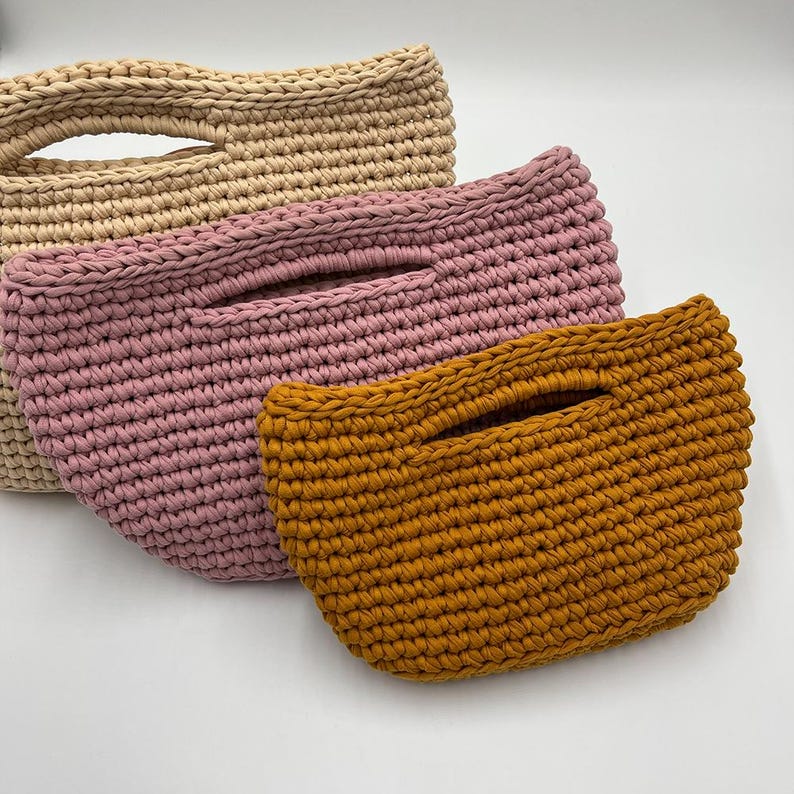 May include: Three handmade crochet bags in beige, pink, and mustard yellow. Each bag features a rectangular shape with a handle opening. The bags are made of thick yarn, showcasing a textured, woven appearance. The bags are displayed on a white surface.