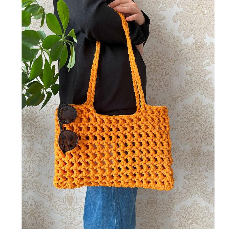 May include: A bright orange crocheted tote bag with a woven pattern. The bag has a wide, flat base and two long straps. Sunglasses are tucked into the bag.