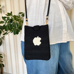 May include: A black crocheted crossbody bag with a white apple logo. The bag has a long strap and is worn over the shoulder.
