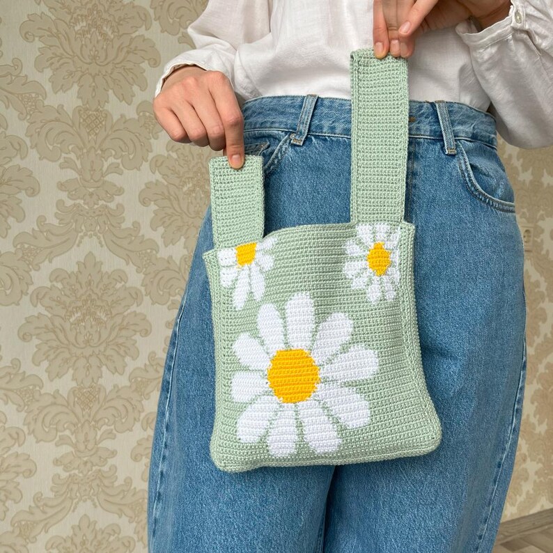 May include: A light green crocheted bag with white and yellow daisy flowers. The bag has two straps and is worn over the shoulder.