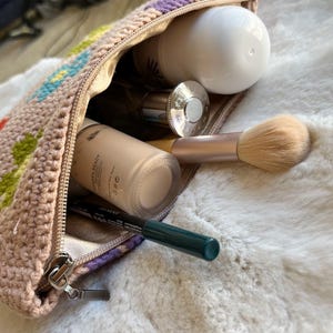 May include: A beige crochet cosmetic bag with a zip, containing makeup items. The bag features colourful floral patterns. Inside are bottles of foundation, perfume, a makeup brush, and an eyeliner pencil.
