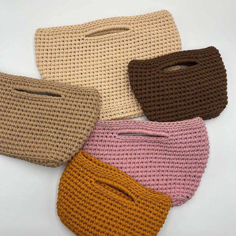 May include: A collection of five crescent-shaped crochet handbags in various solid colors including beige, dark brown, light brown, dusty pink, and mustard yellow. Each bag features a cutout handle integrated into the top edge.