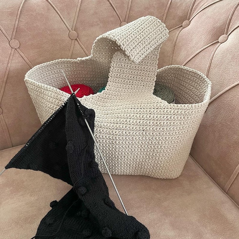 May include: A beige, hand-knitted tote bag with a handle, filled with colorful yarn. A black knitted piece with bobbles is in progress, with knitting needles inserted. The bag is sitting on a pink sofa.