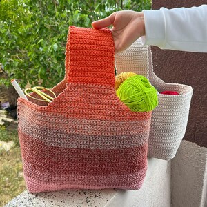 May include: A handmade crochet tote bag in shades of red, orange, and pink, with a matching handle. The bag is filled with yellow and green yarn. A second, smaller beige crochet bag is visible in the background.