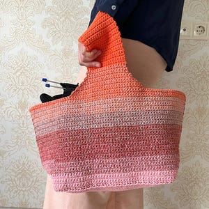 May include: A handmade crochet tote bag in a gradient of colors, from pink to orange. The bag has a wide opening and a sturdy handle. Knitting needles and black yarn are visible inside the bag. The bag is held by a person.