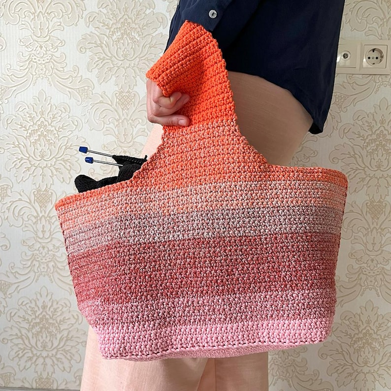 May include: A handmade crochet tote bag in a gradient of colors, from pink to orange. The bag has a wide opening and a sturdy handle. Knitting needles and black yarn are visible inside the bag. The bag is held by a person.