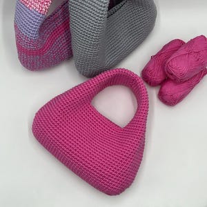 May include: A collection of handmade crochet bags in various colors. The primary bag is a vibrant fuchsia pink with a rounded handle. Other bags are gray and a pink and lavender gradient. Several skeins of pink yarn are also visible.