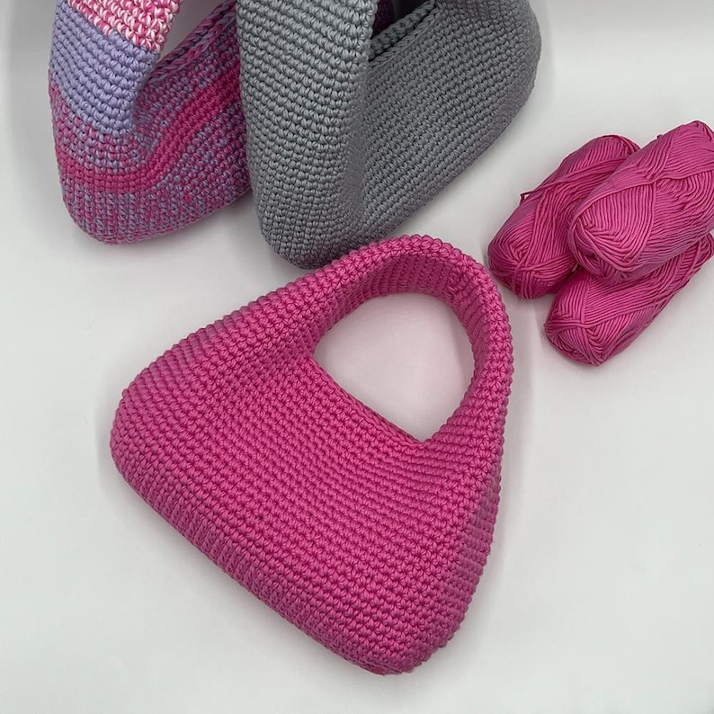 May include: A collection of handmade crochet bags in various colors. The primary bag is a vibrant fuchsia pink with a rounded handle. Other bags are gray and a pink and lavender gradient. Several skeins of pink yarn are also visible.