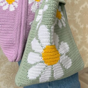 May include: A green crocheted tote bag with a white daisy pattern. The bag has a light purple crocheted section on the top.