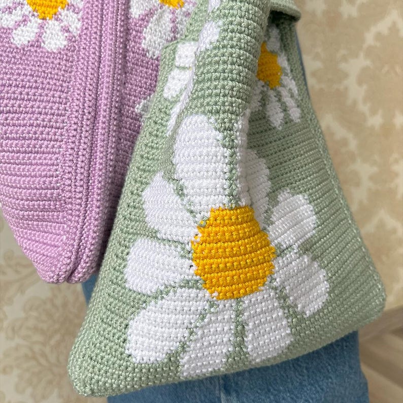 May include: A green crocheted tote bag with a white daisy pattern. The bag has a light purple crocheted section on the top.