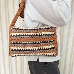Könnte beinhalten: Rechteckige Häkeltasche mit Klappe und langem Riemen. Die Tasche hat horizontale Streifen in Braun, Weiß und Grau. Der Riemen und die Kanten sind einfarbig braun. Handgefertigt. Maße in Zentimetern.
