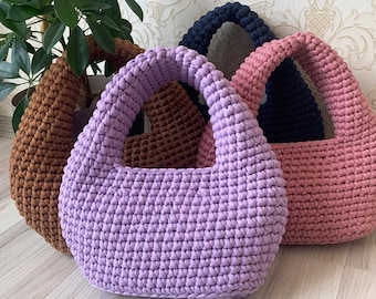 May include: Three handmade crochet handbags in shades of brown, lavender, and pink. Each bag has a rounded shape with a thick, sturdy handle. The bags are displayed against a neutral background, showcasing their texture and color variations.