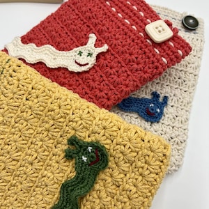 May include: Three crocheted pouches with different colored fabric and embroidered cartoon slugs. The pouches are stacked on top of each other. The top pouch is red, the middle pouch is yellow, and the bottom pouch is beige.