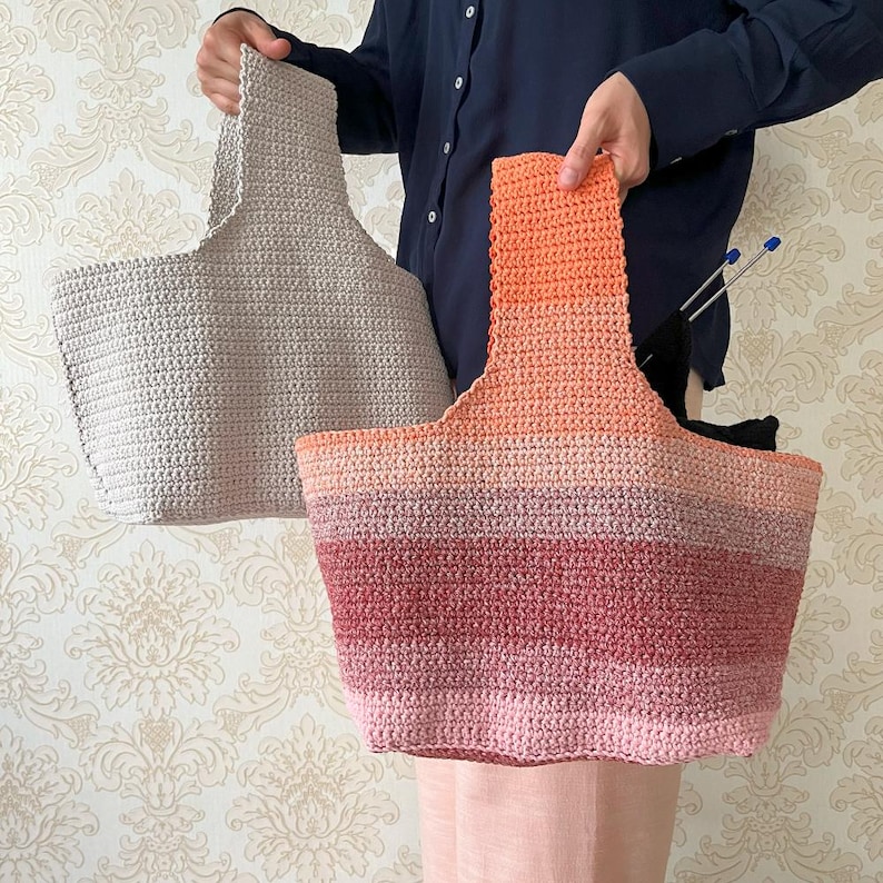 May include: Two handmade crochet tote bags. One is a solid light beige color, and the other is a gradient of pink, red, and orange hues. Both bags have a rectangular base and a wide handle.