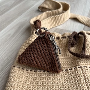 May include: A handmade crochet bag in beige with a brown triangular coin purse attached. The bag has a matching beige strap and brown drawstring closure. The coin purse has a silver zipper and clasp.