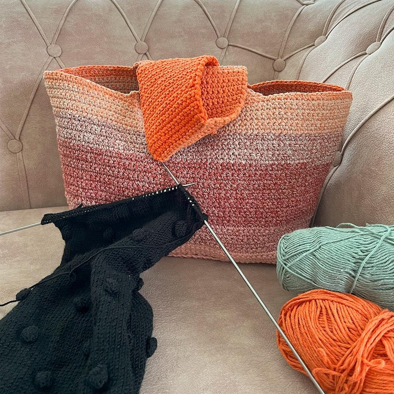 May include: A handmade, crocheted tote bag in shades of orange, peach, and pink, with a matching orange flap. Knitting needles are inserted into a black knitted piece with bobbles. Two balls of yarn, one orange and one teal, are also visible.