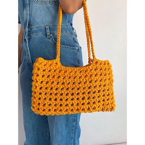 May include: A bright yellow crocheted tote bag with a wide, open top and two long straps. The bag is made of a thick, textured yarn and has a simple, geometric design.