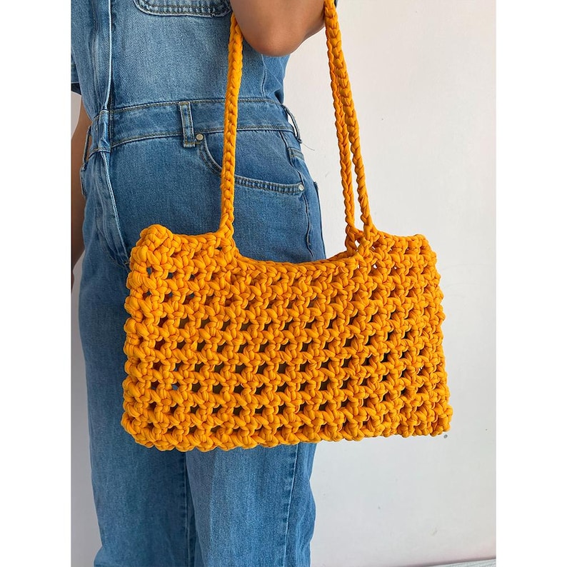 May include: A bright yellow crocheted tote bag with a wide, open top and two long straps. The bag is made of a thick, textured yarn and has a simple, geometric design.