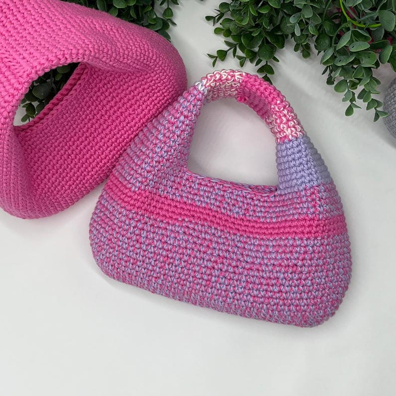 May include: Two handmade crochet bags in shades of pink and purple. One is a large, open-top tote bag, and the other is a small, structured handbag with a handle. The handbag features a striped design and a rounded shape.