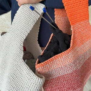 May include: Two handmade crochet bags in the image. One is light gray, and the other is a gradient of orange, peach, and white. Knitting needles with blue tips are visible. The bags contain yarn and black gloves. The bags are made of a soft, textured material.