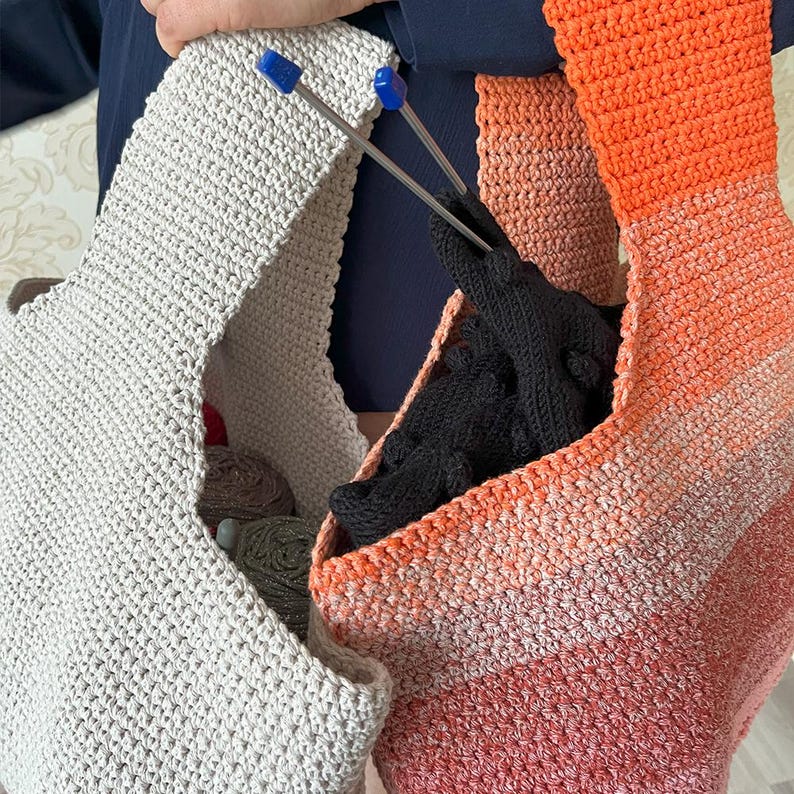 May include: Two handmade crochet bags in the image. One is light gray, and the other is a gradient of orange, peach, and white. Knitting needles with blue tips are visible. The bags contain yarn and black gloves. The bags are made of a soft, textured material.