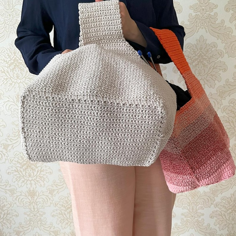 May include: Two handmade crochet bags. One is a light beige tote bag with a rectangular base and a handle. The other is a shoulder bag with a gradient of pink, orange, and red. Both bags are made with a tight crochet stitch.