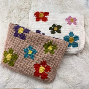 May include: Two crochet pouches with colourful flower designs. One pouch is beige with purple, blue, green, and red flowers. The other is white with red, pink, green, and blue flowers. The pouches are on a white, fluffy surface.