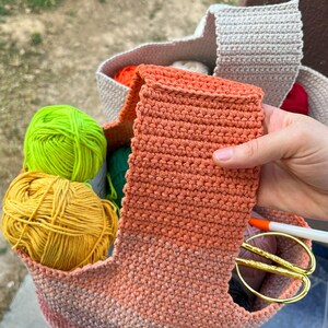 May include: A crocheted tote bag filled with colorful yarn balls in shades of yellow, green, and orange. An orange crocheted item is held up, with scissors and a marker visible inside the bag. The bag is beige and orange.