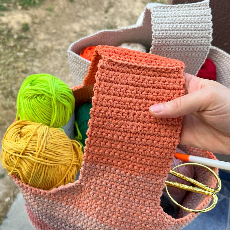 May include: A crocheted tote bag filled with colorful yarn balls in shades of yellow, green, and orange. An orange crocheted item is held up, with scissors and a marker visible inside the bag. The bag is beige and orange.