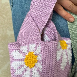 May include: A crocheted tote bag with a purple base and white daisy designs. The daisies have yellow centers.