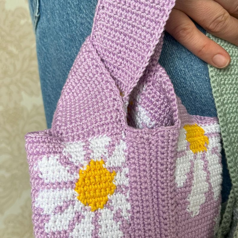 May include: A crocheted tote bag with a purple base and white daisy designs. The daisies have yellow centers.