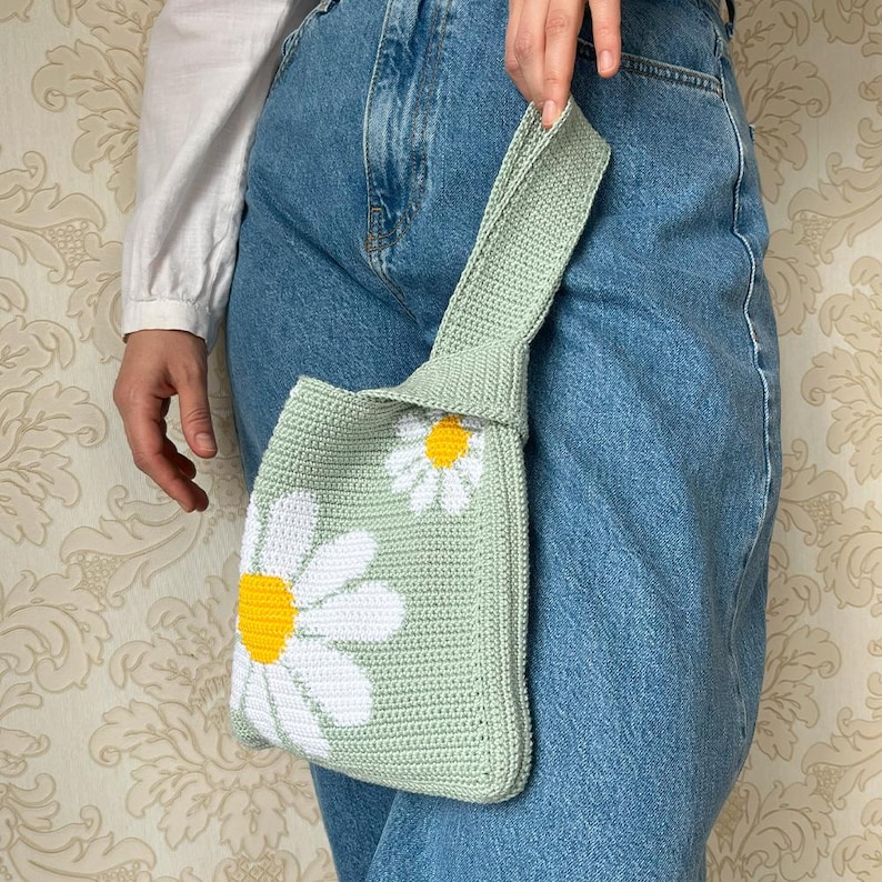 May include: A light green crocheted tote bag with white daisy flower designs. The bag has a long strap and is being held by a person wearing blue jeans.