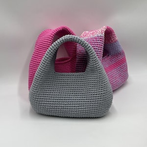 May include: Three handmade crochet bags in shades of pink and gray. The front-most bag is gray with a rounded shape and a handle. The other two bags are in shades of pink, with one solid and the other with a striped pattern.