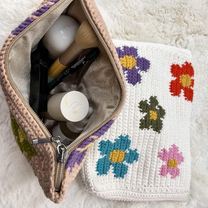 May include: A crocheted makeup bag with a zip, filled with beauty products, next to a matching white bag with colourful floral designs. The open bag is beige with purple and green accents.