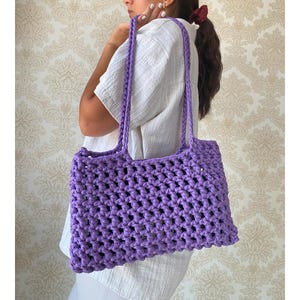 May include: A purple crocheted tote bag with a large open weave pattern. The bag has two straps and is being held by a person.