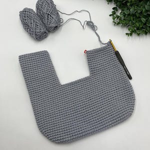 May include: A work-in-progress crochet project in gray yarn. The piece is a U-shaped design, with two balls of yarn and a crochet hook visible. A red stitch marker is placed on the edge of the project. The background is white with a touch of greenery.